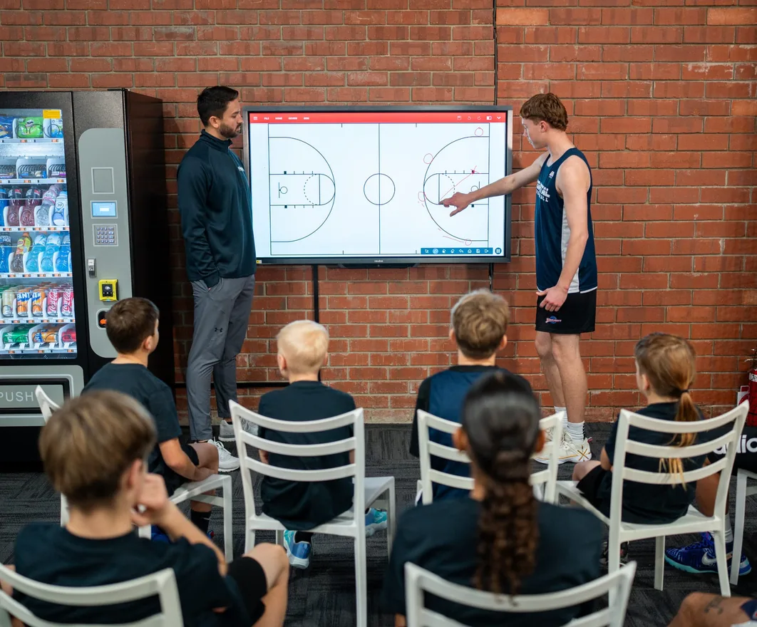 A coach and player working through a play on a digital whiteboard while a junior squad watches